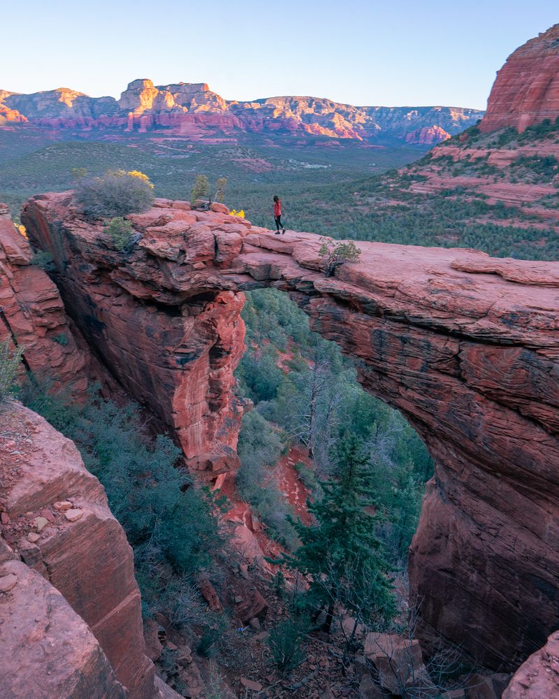 A hiker standing on Devil's Bridge, a red sandstone arch in Sedona, with red rock mesas behind